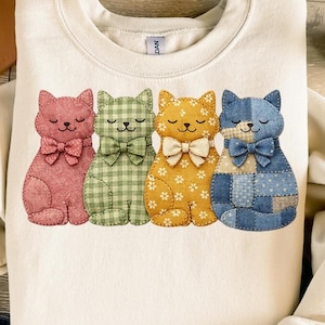 May include: Cream-colored sweatshirt featuring four appliqued cats in pink, green gingham, yellow floral, and blue patchwork patterns. Each cat has a bow tie. The design is centered on the front of the shirt.
