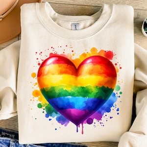 Puede incluir: Sudadera de color crema con un diseño de corazón grande y colorido. El corazón está pintado con un arcoíris de colores, con tonos rojos, naranjas, amarillos, verdes, azules y morados. Salpicaduras de pintura rodean el corazón, añadiendo un toque vibrante.