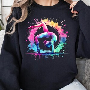 May include: Black sweatshirt with a colorful watercolor design of a gymnast performing a handstand on a ring. The design features splashes of pink, blue, green, and yellow.