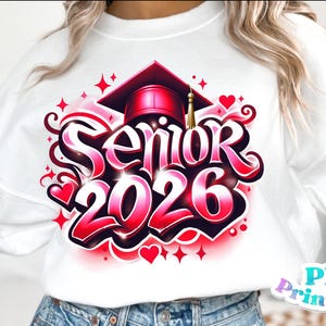 K&ouml;nnte beinhalten: Wei&szlig;es Sweatshirt mit einem farbenfrohen Design f&uuml;r den Abschlussjahrgang 2026. Das Design zeigt die Worte "Senior 2026" in einer fetten Schrift, eine Abschlusskappe, Herzen und Sterne. Das Design ist in Rot- und Rosat&ouml;nen gehalten.