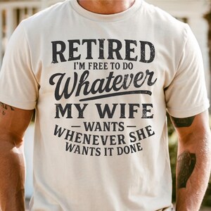 May include: A cream-coloured t-shirt featuring the text "RETIRED I'M FREE TO DO Whatever MY WIFE WANTS WHENEVER SHE WANTS IT DONE" in a distressed black font. The shirt has a crew neck and short sleeves.