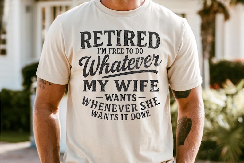 May include: A cream-coloured t-shirt featuring the text "RETIRED I'M FREE TO DO Whatever MY WIFE WANTS WHENEVER SHE WANTS IT DONE" in a distressed black font. The shirt has a crew neck and short sleeves.