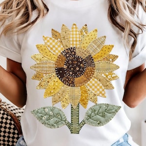 May include: White t-shirt featuring a large, detailed sunflower design. The sunflower has yellow and brown patchwork petals, a brown center, and green leaves. The design is centered on the shirt.