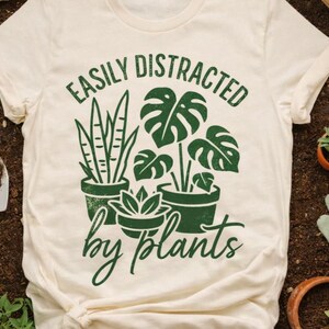 以下が含まれることがあります： 緑色の「EASILY DISTRACTED by plants」の文字が入った、クリーム色のTシャツ。 蛇の植物やモンステラなど、鉢植えの植物のグラフィックが特徴です。 庭いじりのテーマ。