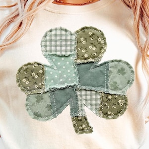 以下が含まれることがあります： クリーム色のスウェットシャツ。大きなパッチワークのクローバーのデザインが特徴です。クローバーは、花柄、水玉模様、ギンガムチェックなど、さまざまな緑色の生地で構成されています。クローバーの端はほつれており、素朴な印象を与えます。