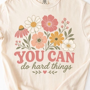May include: Cream-colored t-shirt featuring a floral design with pink, peach, yellow, and white flowers. The text "YOU CAN do hard things" is in pink and green. The shirt has rolled-up sleeves.