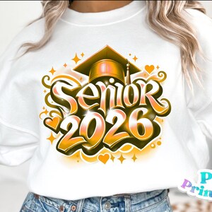 K&ouml;nnte beinhalten: Wei&szlig;es Sweatshirt mit einem Abschluss-Design. Das Design zeigt den Text "Senior 2026" in einer stilisierten Schriftart, eine Abschlusskappe, Sterne und Herzen in Gold- und Oranget&ouml;nen. Das Sweatshirt ist ein l&auml;ssiges Kleidungsst&uuml;ck.