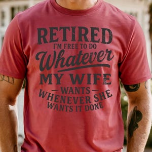 May include: A maroon t-shirt with the text "RETIRED I'M FREE TO DO Whatever MY WIFE wants WHENEVER SHE WANTS IT DONE" in black lettering. The shirt is short-sleeved and appears to be made of cotton.