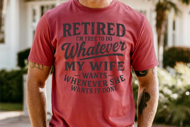 May include: A maroon t-shirt with the text "RETIRED I'M FREE TO DO Whatever MY WIFE wants WHENEVER SHE WANTS IT DONE" in black lettering. The shirt is short-sleeved and appears to be made of cotton.