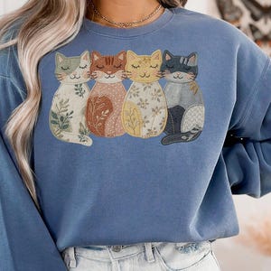 Pu&ograve; includere: Una felpa blu con un ricamo di quattro gatti stilizzati. Ogni gatto ha un motivo unico, tra cui disegni floreali e patchwork, nelle tonalit&agrave; del verde, marrone, giallo e grigio. La felpa &egrave; di colore blu unito.