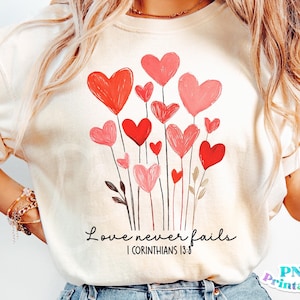 Puede incluir: Camiseta blanca con un diseño de corazones rojos y rosas en tallos. El texto "El amor nunca falla 1 Corintios 13:8" está impreso debajo de los corazones.