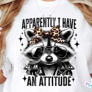 May include: White t-shirt featuring a black and white illustration of a raccoon wearing sunglasses and a leopard print bow. The text "APPARENTLY I HAVE AN ATTITUDE" is printed on the shirt.