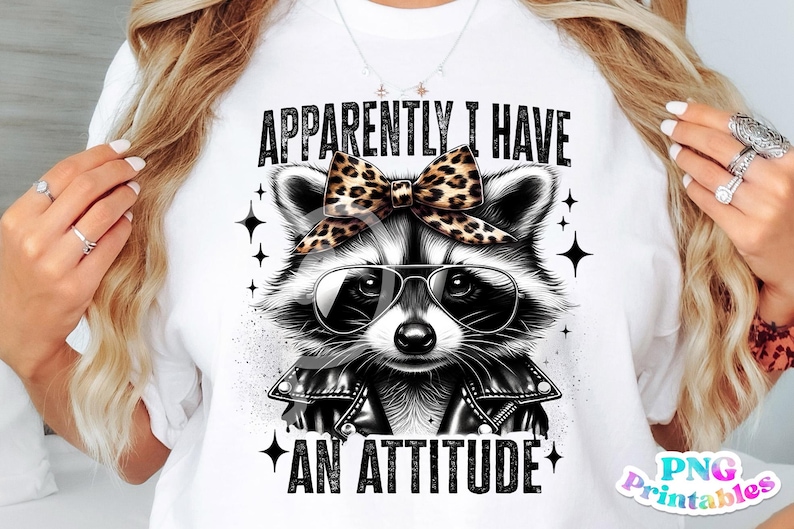 May include: White t-shirt featuring a black and white illustration of a raccoon wearing sunglasses and a leopard print bow. The text "APPARENTLY I HAVE AN ATTITUDE" is printed on the shirt.