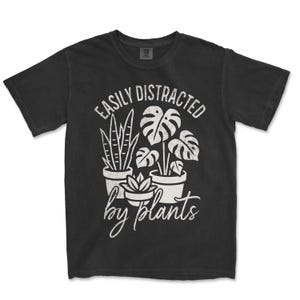 以下が含まれることがあります： 黒いTシャツ。白いグラフィックで鉢植えの植物と「EASILY DISTRACTED by plants」の文字。デザインには、サンスベリアとモンステラの葉が含まれています。