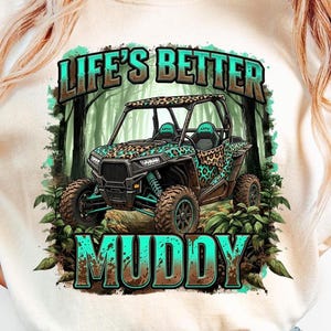 Könnte beinhalten: Ein cremefarbenes T-Shirt mit einem Grafikdesign eines UTV mit Leopardenmuster und dem Text "LIFE'S BETTER MUDDY" in Türkis und Braun. Das UTV ist von einer Waldszene umgeben.