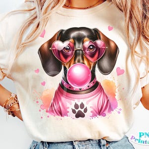 May include: A cream-colored t-shirt featuring a dachshund wearing heart-shaped sunglasses and a pink sweater. The dog has a pink bubble gum bubble and a paw print on the sweater. Pink hearts and watercolor splashes surround the dog.