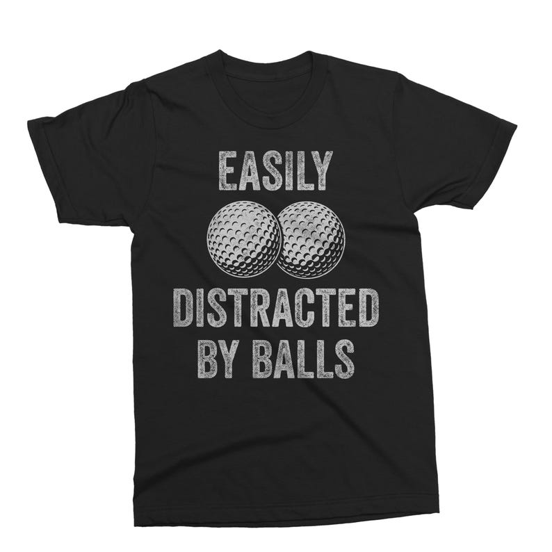 Puede incluir: Camiseta negra con el texto "EASILY DISTRACTED BY BALLS" en letras blancas desgastadas. Dos pelotas de golf est&aacute;n centradas encima del texto. La camiseta es de cuello redondo.