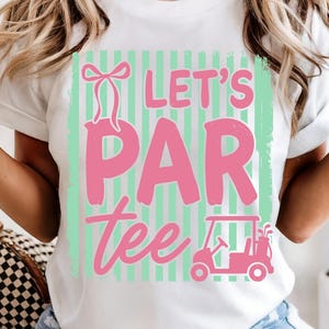 May include: White t-shirt featuring a pink and green graphic. The design reads "Let's Par Tee" with a golf cart illustration. The shirt has a casual, sporty theme.