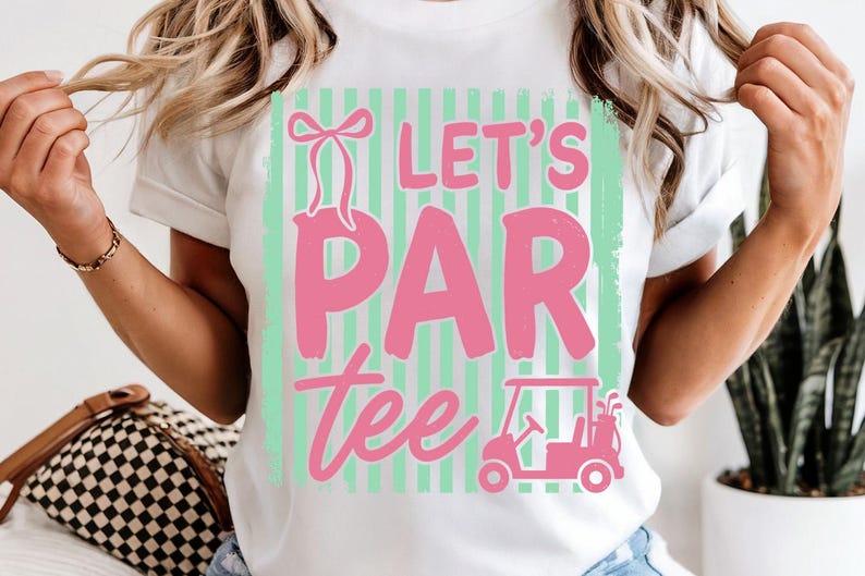 May include: White t-shirt featuring a pink and green graphic. The design reads "Let's Par Tee" with a golf cart illustration. The shirt has a casual, sporty theme.
