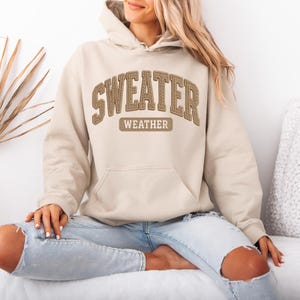 May include: A beige hoodie with the words "SWEATER WEATHER" in brown stitched lettering across the chest. The hoodie has a front pocket and a hood. The person is wearing light blue jeans.