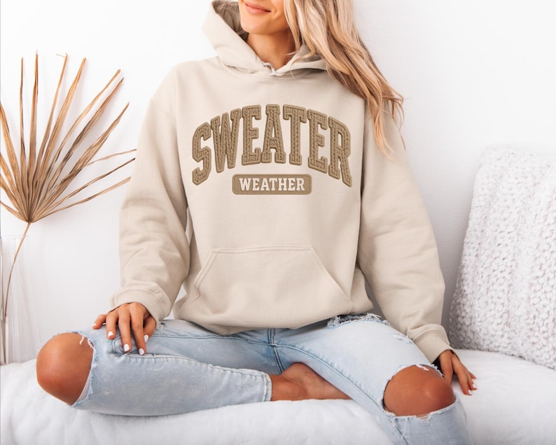 May include: A beige hoodie with the words "SWEATER WEATHER" in brown stitched lettering across the chest. The hoodie has a front pocket and a hood. The person is wearing light blue jeans.