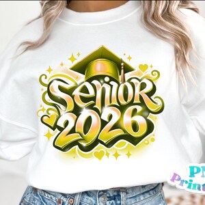K&ouml;nnte beinhalten: Wei&szlig;es Sweatshirt mit einer Abschlusskappe-Grafik und dem Text "Senior 2026" in einem stilisierten gelb-gr&uuml;nen Design. Das Design enth&auml;lt Herzen und Sterne. Ein Bekleidungsartikel zum Thema Abschluss.