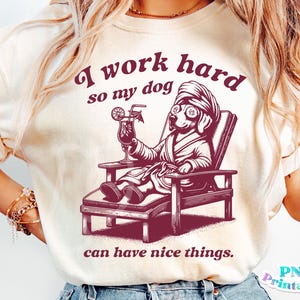 Może przedstawiać: Biały t-shirt z r&oacute;żowym nadrukiem psa w szlafroku i okularach przeciwsłonecznych, trzymającego drinka. Tekst na koszulce brzmi: "Ciężko pracuję, żeby m&oacute;j pies m&oacute;gł mieć fajne rzeczy."