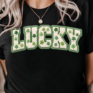 以下が含まれることがあります： 「LUCKY」の文字が緑と白のデザインで描かれた黒いTシャツ。文字は緑のクローバーで埋められ、緑で縁取られています。カジュアルなスタイルのTシャツです。