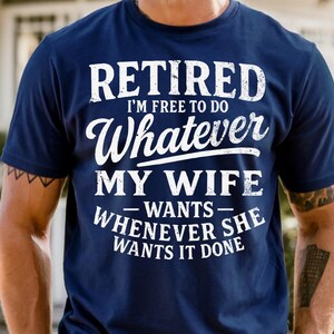 May include: Navy blue t-shirt with white text that reads "RETIRED I'M FREE TO DO Whatever MY WIFE WANTS WHENEVER SHE WANTS IT DONE". The shirt is a crew neck and has short sleeves.