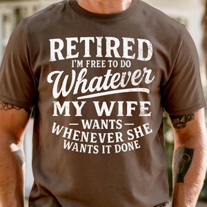 May include: A brown t-shirt with the text "RETIRED I'M FREE TO DO Whatever MY WIFE WANTS WHENEVER SHE WANTS IT DONE" in white lettering. The shirt is short-sleeved.