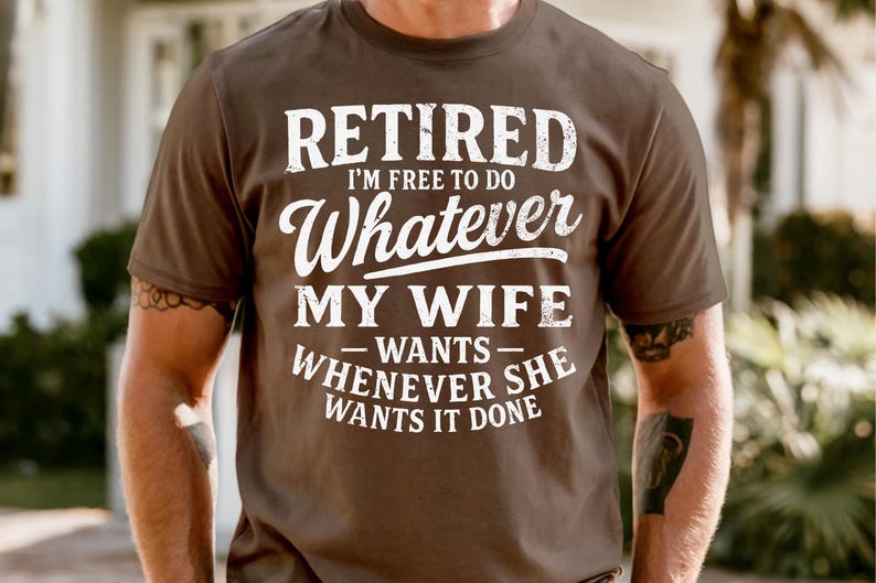 May include: A brown t-shirt with the text "RETIRED I'M FREE TO DO Whatever MY WIFE WANTS WHENEVER SHE WANTS IT DONE" in white lettering. The shirt is short-sleeved.