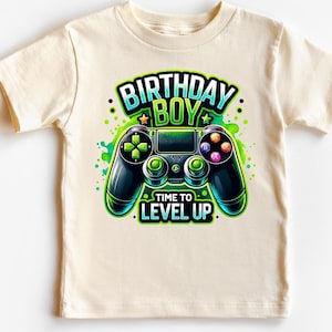 May include: A light beige t-shirt with a green and black video game controller graphic. The text "Birthday Boy" is above the controller and "Time to Level Up" is below the controller.