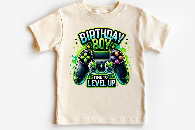 May include: A light beige t-shirt with a green and black video game controller graphic. The text "Birthday Boy" is above the controller and "Time to Level Up" is below the controller.