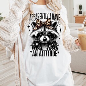 May include: White t-shirt featuring a black and white illustration of a raccoon wearing sunglasses and a bow. The text "Apparently I have an attitude" is printed on the shirt.