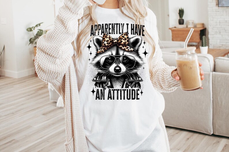 May include: White t-shirt featuring a black and white illustration of a raccoon wearing sunglasses and a bow. The text "Apparently I have an attitude" is printed on the shirt.