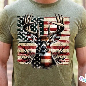 May include: Olive green t-shirt featuring a deer head design with antlers, superimposed over an American flag. The deer's face is detailed with the flag's stars and stripes. The shirt is a casual wear item.