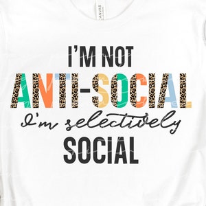 May include: White t-shirt with the text "I'm not anti-social I'm selectively social" in black. The word "anti-social" is in colorful letters with a leopard print pattern.