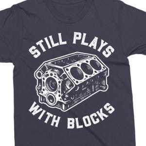 Peut inclure: T-shirt bleu marine avec un motif blanc d'un bloc moteur et le texte "STILL PLAYS WITH BLOCKS". Le design est une illustration détaillée d'un moteur, parfait pour les passionnés de voitures.