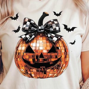May include: A white t-shirt with a gold disco ball pumpkin with a black and white checkered bow. Bats are scattered around the pumpkin.