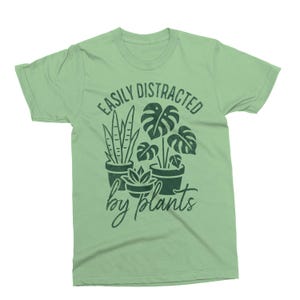 以下が含まれることがあります： 薄い緑色のTシャツ。テキスト「EASILY DISTRACTED by plants」は濃い緑色。グラフィックは、多肉植物、大きな葉を持つ植物、細長い葉を持つ植物の3つの鉢植えの植物を描いています。