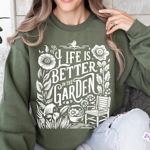 May include: A green sweatshirt with a white graphic design that says "Life is Better in the Garden". The design features flowers, leaves, a bird, a chair, and a pot.