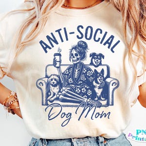 Może przedstawiać: Biały T-shirt z niebieskim nadrukiem graficznym, na kt&oacute;rym widnieje napis "Anti-Social Dog Mom". Grafika przedstawia szkielet trzymający kubek kawy i siedzący na kanapie z trzema psami.