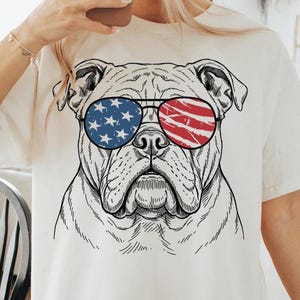 Puede incluir: Camiseta color crema con una ilustración en blanco y negro de un bulldog con gafas de sol. Las gafas de sol tienen un diseño patriótico, con estrellas azules en una lente y rayas rojas en la otra.