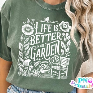 Può includere: Una maglietta verde con un design grafico bianco che dice "Life is better in the garden". Il design presenta fiori, foglie, un uccello, una sedia e un vaso.