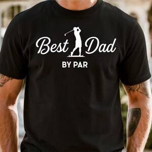 K&ouml;nnte beinhalten: Schwarzes T-Shirt mit dem Text "Best Dad" in Schreibschrift, begleitet von einer Golfspieler-Silhouette. Der Schriftzug "BY PAR" steht darunter. Das T-Shirt hat kurze &Auml;rmel.