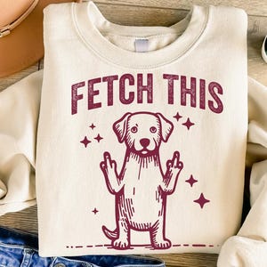 May include: A cream-colored sweatshirt with a maroon graphic of a dog giving the middle finger with the text "FETCH THIS" above it.