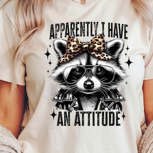May include: A white t-shirt featuring a black and white illustration of a raccoon wearing sunglasses and a leopard print bow. The text "Apparently I have an attitude" is printed on the shirt.