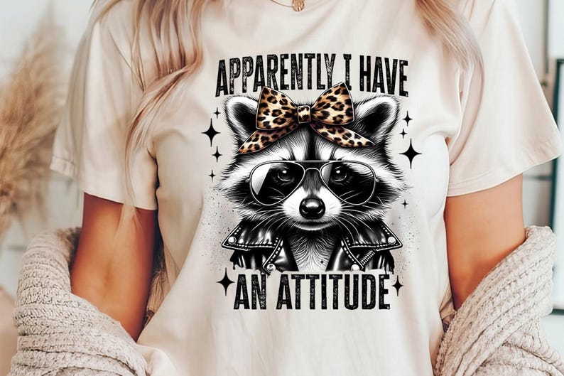 May include: A white t-shirt featuring a black and white illustration of a raccoon wearing sunglasses and a leopard print bow. The text "Apparently I have an attitude" is printed on the shirt.