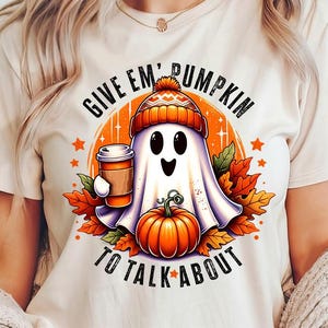 May include: A white t-shirt with a graphic of a ghost holding a cup of coffee and a pumpkin. The text "Give 'em Pumpkin to Talk About" is printed on the shirt.
