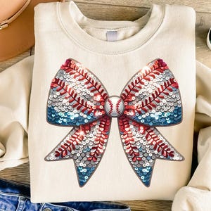 May include: A white sweatshirt with a large sequined bow in red, white, and blue colors. The bow is shaped like a baseball with red stitching and a white baseball in the center.